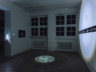 Schummerig beleuchteter Galerieraum mit kreisförmig auf dem Boden angeordneten glasähnlichen Objekten, einem Scheinwerfer an der rechten Wand und an den Wänden ausgestellten Kunstwerken.