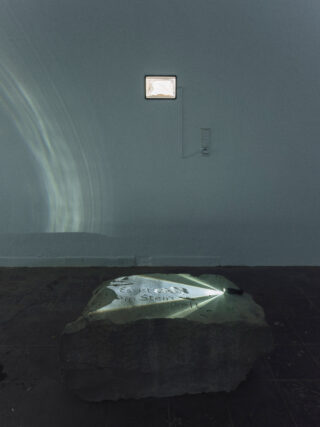 Auf dem dunklen Boden liegt eine raue Steinplatte mit einem von Licht beleuchteten Text; an der leeren Wand dahinter sind ein kleines Fenster und eine Steckdose zu sehen.