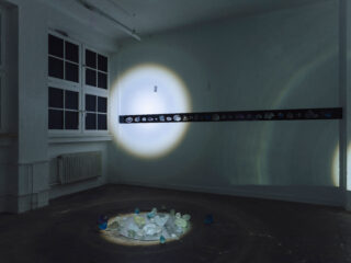 Ein schwach beleuchteter Raum mit einem Scheinwerfer, der einen Kreis aus verschiedenen durchsichtigen Steinen auf dem Boden und eine horizontale Reihe von Steinen an der Wand beleuchtet.