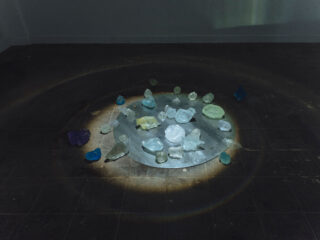 Verschiedene durchscheinende farbige Steine sind in einem kreisförmigen Muster auf einer runden Metallfläche angeordnet, wobei ein schwacher Lichtring auf den dunklen Fliesenboden um sie herum geworfen wird.