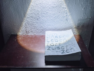 Ein strukturiertes Buch mit geprägtem deutschen Text liegt auf einem dunklen Holztisch unter einem gebündelten Lichtstrahl vor einer rauen weißen Wand.
