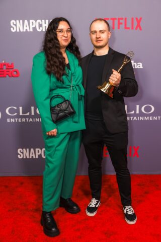Zwei Personen stehen auf einem roten Teppich; eine hält eine Trophäe. Sie posieren vor einer Kulisse mit Logos wie Netflix, Clio Entertainment und Snapchat.