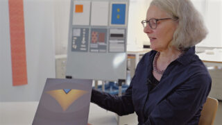 Eine Frau mit grauem Haar und Brille hält ein Blatt Papier mit geometrischer Kunst; im Hintergrund steht eine Schautafel mit Designmustern.