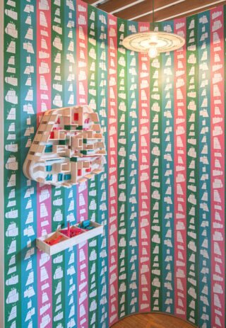 Eine geometrische Wandinstallation mit Regalen und farbigen Blöcken ist an einer Eckwand angebracht, die mit einer sich wiederholenden rot, grün und weiß gemusterten Tapete bedeckt ist. Darüber hängt eine einzelne Deckenleuchte.