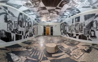 Ein Raum, dessen Boden, Wände und Decke mit Schwarz-Weiß-Collagen mit Texten und Porträts bedeckt sind; in der Mitte steht ein Baumstumpf.