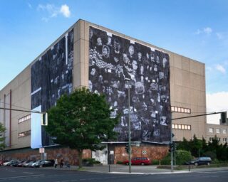 Ein großes beigefarbenes Gebäude mit einer Wandmalerei aus schwarz-weißen Porträts an der Eckwand; im Vordergrund stehen geparkte Autos und ein Baum.