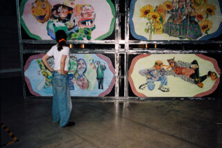 Eine Person in weißem Hemd und Jeans steht vor vier farbenfrohen, surrealen Gemälden, die an einer Wand in einem Innenraum hängen.