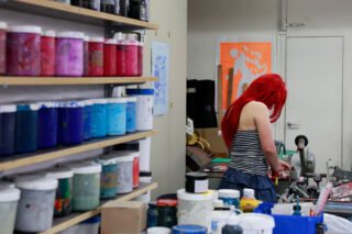 Eine Person mit langen roten Haaren arbeitet an einem Tisch in einem Kunstatelier, umgeben von Regalen mit bunten Farbdosen und Kunstzubehör.