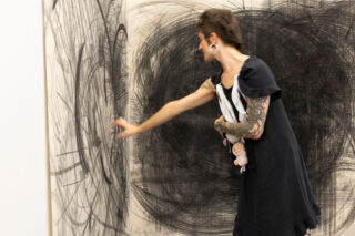 Eine Person mit Tätowierungen und Ohrringen zeichnet große kreisförmige Muster auf eine mit schwarzem und grauem Gekritzel bedeckte Wand und hält ein Stofftier in einem Arm.