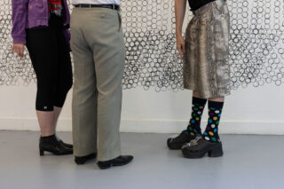 Drei Personen stehen in einem Innenraum, von der Taille abwärts sichtbar, in unterschiedlicher Kleidung, darunter gemusterte Röcke, gepunktete Socken und Kleiderschuhe, vor einer Wand mit kreisförmiger Drahtkunst.