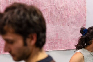 Zwei Personen stehen vor einer Wand mit einem großen, strukturierten rosa Kunstwerk; beide Personen sind teilweise unscharf und blicken in unterschiedliche Richtungen.