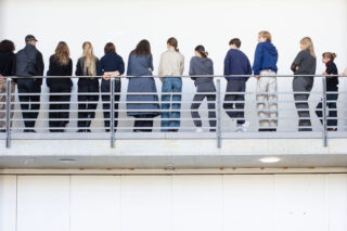 Eine Gruppe von Menschen steht in einer Reihe auf einem Balkon und blickt mit dem Rücken zur Kamera auf eine weiße Wand.