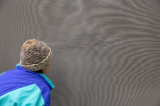Eine Person in einer blauen und blaugrünen Jacke mit einer Strickmütze betrachtet eine neblige oder beschlagene Glasfläche genau.