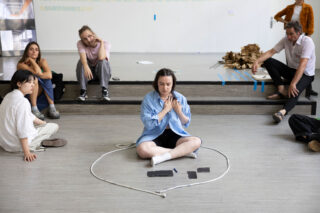 Eine Gruppe von Menschen sitzt um eine Frau herum, die im Schneidersitz in einem Seilkreis auf dem Boden sitzt und vor der verschiedene Gegenstände platziert sind.