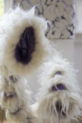 Nahaufnahme eines weißen, pelzigen skulpturalen Objekts mit lila Innenfutter, das in einem Raum mit gemustertem Hintergrund ausgestellt ist.