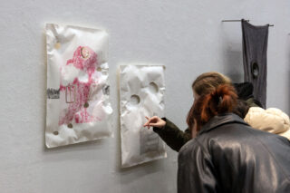 Zwei Personen betrachten Papierkunstwerke mit Löchern, die an einer Galeriewand ausgestellt sind; ein Kunstwerk hat ein rosa abstraktes Design, das andere wirkt eher minimalistisch.