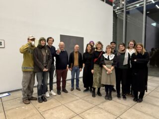 Eine Gruppe von dreizehn Personen steht in einem Innenraum vor einer weißen Wand und posiert für ein Foto. Einige lächeln, und eine Person hält ein Getränk hoch.