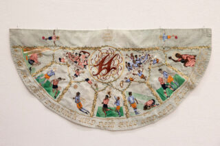 Ein halbkreisförmiges besticktes Textil zeigt verschiedene Fußballszenen, Spieler und Trophäen, mit dekorativen Goldelementen und einem Monogramm in der Mitte, das an einer weißen Wand hängt.