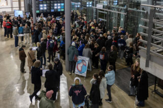 Eine große Menschenmenge steht in der Lobby eines modernen Gebäudes, einige in der Nähe eines Plakats mit einem Vogel, während andere in Schlangen warten oder sich in kleinen Gruppen zusammenfinden.