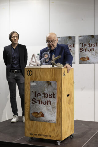 Zwei Männer stehen auf einer Bühne; einer spricht an einem hölzernen Podium mit einem Mikrofon, während der andere in der Nähe steht. Im Hintergrund sind Plakate mit der Aufschrift 