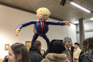 Eine Gruppe von Menschen steht in der Nähe einer großen schwebenden Figur mit übertriebenen Gesichtszügen, gekleidet in einen blauen Anzug und eine rote Krawatte, die in einem überdachten Galerieraum ausgestellt ist.