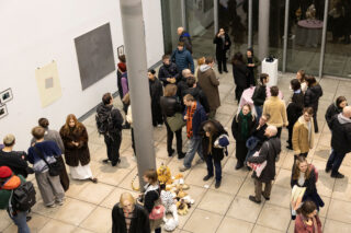 Eine Gruppe von Menschen besucht eine Kunstausstellung in einem Galerieraum; einige betrachten Kunstwerke an den Wänden, während andere sich um Plüschtiere auf dem Boden versammeln.