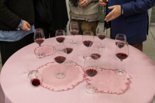 Ein runder Tisch mit mehreren Rotweingläsern, von denen einige Ringflecken auf der rosa Tischdecke hinterlassen, und drei Personen, die in der Nähe stehen. Ein Glas ist umgekippt.