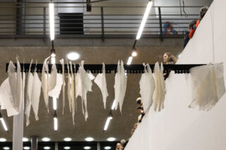 Blätter aus durchscheinendem Papier hängen an einer Metallstange in einem Innenraum und werden von einem Balkon im Obergeschoss aus beobachtet.