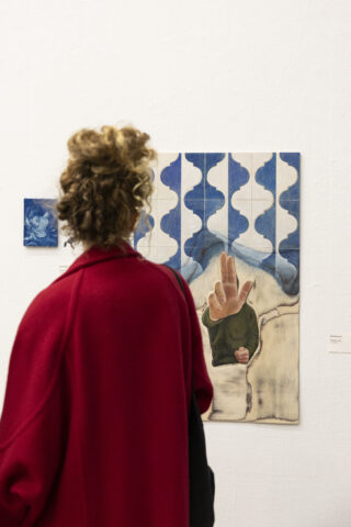 Eine Person in einem roten Mantel steht vor zwei abstrakten Gemälden an einer weißen Wand und betrachtet die Kunstwerke genau.