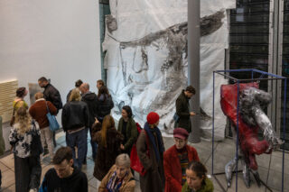Eine Gruppe von Menschen versammelt sich in einer Kunstgalerie und betrachtet Skulpturen und ein großes abstraktes Wandgemälde in Schwarz-Weiß-Tönen.