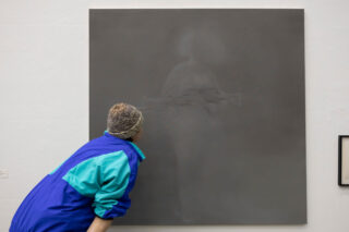 Eine Person in einer bunten Jacke betrachtet aufmerksam ein großes, graues, abstraktes Gemälde mit einer schwachen, undeutlichen Figur darauf, das an einer weißen Wand hängt.