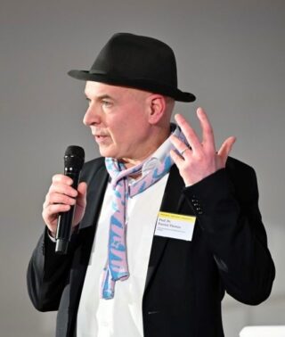 Ein Mann, der einen schwarzen Hut und einen gemusterten Schal trägt, spricht während einer Präsentation in ein Mikrofon und hält dabei seine Hand mit drei ausgestreckten Fingern hoch.