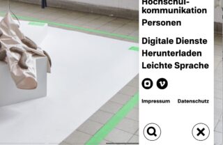 Ein weißes, rechteckiges Podest, auf dem ein beiger Stoff drapiert ist und das mit grünem Klebeband eingefasst ist, steht auf einem gefliesten Boden neben dem Navigationsmenü einer Website.