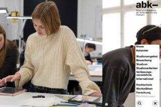 Eine Frau in einem cremefarbenen Pullover beugt sich über einen Tisch und arbeitet mit Papieren und Kunstzubehör in einem Klassenzimmer der Staatlichen Akademie der Bildenden Künste Stuttgart.