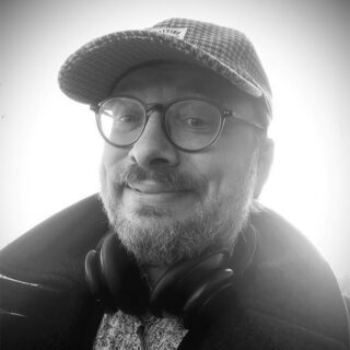 Eine Person mit Brille, karierter Mütze und Kopfhörern um den Hals lächelt auf einem Schwarz-Weiß-Foto in die Kamera.