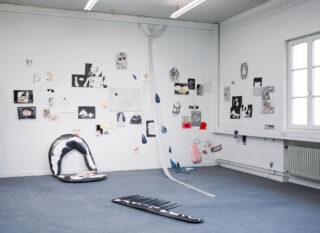 Kunstinstallation in einem weißen Raum mit Mixed-Media-Kunstwerken an der Wand, einem aufgehängten Netz mit Ketten und Tropfen und skulpturalen Stücken auf dem Boden.