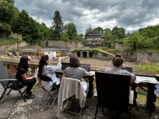 Vier Personen sitzen mit Laptops und Getränken an einem Tisch und blicken auf eine archäologische Ausgrabungsstätte mit Baumaschinen und einem teilweise bewölkten Himmel im Hintergrund.