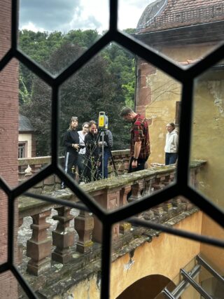 Eine Gruppe von fünf Personen steht auf einem alten Steinbalkon mit Vermessungsgeräten, die durch ein Fenster mit einem geometrischen Gittermuster betrachtet werden.