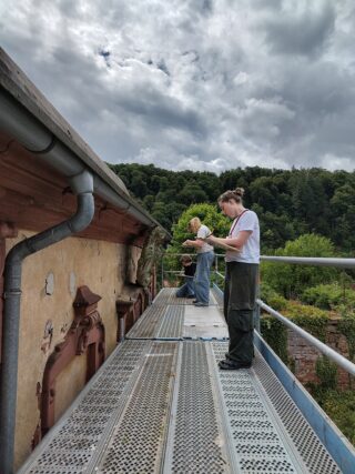 Zwei Personen stehen auf einem Metallgerüst neben einem alten Gebäude und machen sich Notizen oder machen Beobachtungen, während im Hintergrund Bäume und ein bewölkter Himmel zu sehen sind.