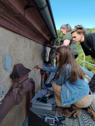 Drei Personen untersuchen die Außenwand eines alten Gebäudes und nehmen mit wissenschaftlichen Geräten Proben.