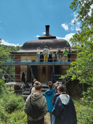 Eine Gruppe von Menschen steht auf einem Gerüst um ein kleines Gebäude herum, während andere unten, umgeben von Grün unter einem teilweise bewölkten Himmel, beobachten.