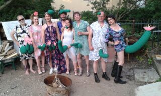 Eine Gruppe von acht Erwachsenen posiert im Freien in bunten Kleidern, einige halten oder tragen grüne Kostüme, die Pflanzen mit rosa Blüten ähneln. Im Vordergrund ist eine Feuerstelle zu sehen.