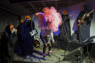 Eine Band tritt auf der Bühne auf; die Mitglieder tragen schwarze Gewänder und Emoji-Gesichtsmasken. Der Frontmann trägt ein rosa Kostüm, einen großen Kopfschmuck und hält ein Schild mit der Aufschrift 