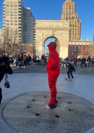 Eine mit roter Schnur umwickelte Person steht auf einem Gitter vor dem Washington Square Arch, während im Hintergrund Menschen sitzen und ein Skateboarder zu sehen ist.