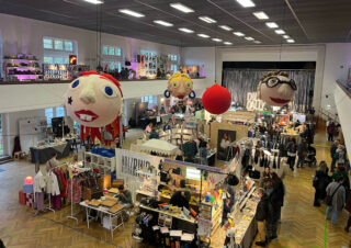 Indoor-Kunsthandwerksmarkt mit verschiedenen Verkaufsständen, Einkäufern und großen dekorativen Puppenköpfen, die in einer gut beleuchteten Halle von der Decke hängen.