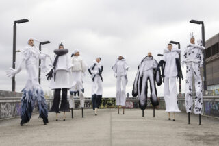 Acht Personen auf Stelzen in avantgardistischen weißen Kostümen mit übertriebenen Formen und Akzenten, die an einem bewölkten Tag im Freien stehen, mit Graffiti bedeckten Wänden im Hintergrund.