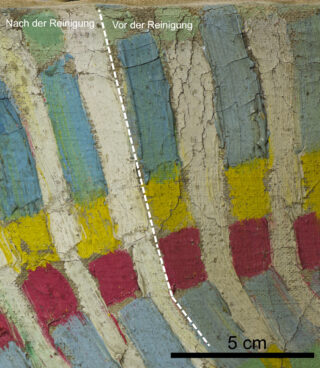 Nahaufnahme einer bemalten Oberfläche mit einer gestrichelten Linie, die gereinigte (links) und ungereinigte (rechts) Bereiche voneinander trennt, mit sichtbaren Farbunterschieden und einem 5-cm-Skalierungsbalken am unteren Rand.