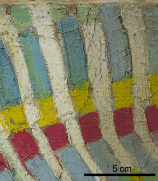 Nahaufnahme einer bemalten Oberfläche mit blauen, weißen, gelben und roten vertikalen Streifen. Die Farbe ist rissig, was Textur und Alter erkennen lässt. Schwarzer Maßstabsbalken von 5 cm unten rechts.