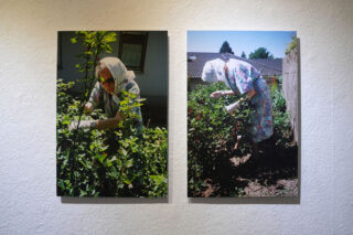 Auf einer weißen Wand sind zwei Fotos einer älteren Frau mit Kopftuch und Handschuhen zu sehen, die sich in einem Garten um Sträucher kümmert.