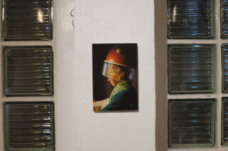 Ein Foto einer Person im Profil unter einem alten orangefarbenen Salon-Haartrockner ist an eine weiße Wand zwischen Glasblockfenstern geklebt.